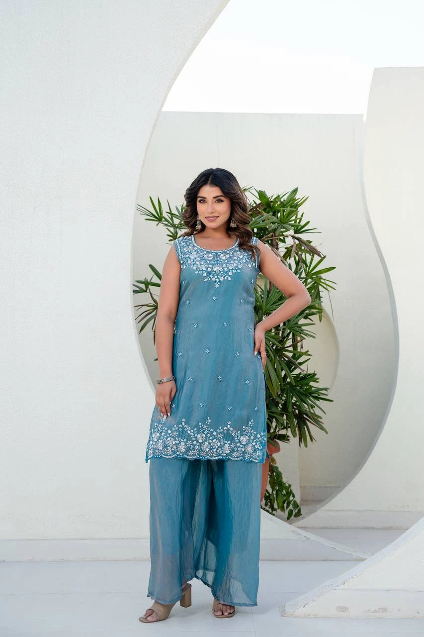 Woman wearing a blue embroidered outfit standing in front of a white arch with greenery.